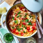A pot of unstuffed cabbage roll soup with tomatoes, rice, and fresh herbs sits on a wooden table beside a slice of bread, utensils, and a small bowl of chopped parsley.