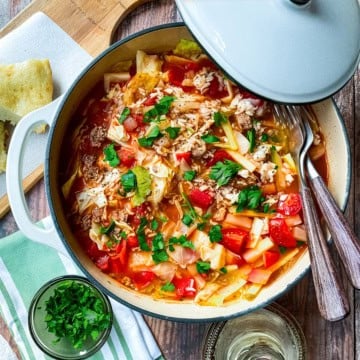 A pot of unstuffed cabbage roll soup with tomatoes, rice, and fresh herbs sits on a wooden table beside a slice of bread, utensils, and a small bowl of chopped parsley.
