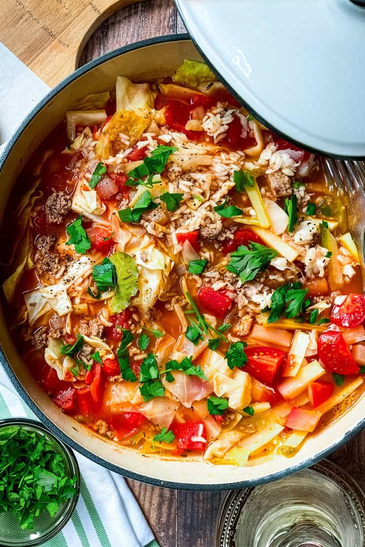 A pot of unstuffed cabbage roll soup with tomatoes, rice, and fresh herbs sits on a wooden table beside a slice of bread, utensils, and a small bowl of chopped parsley.