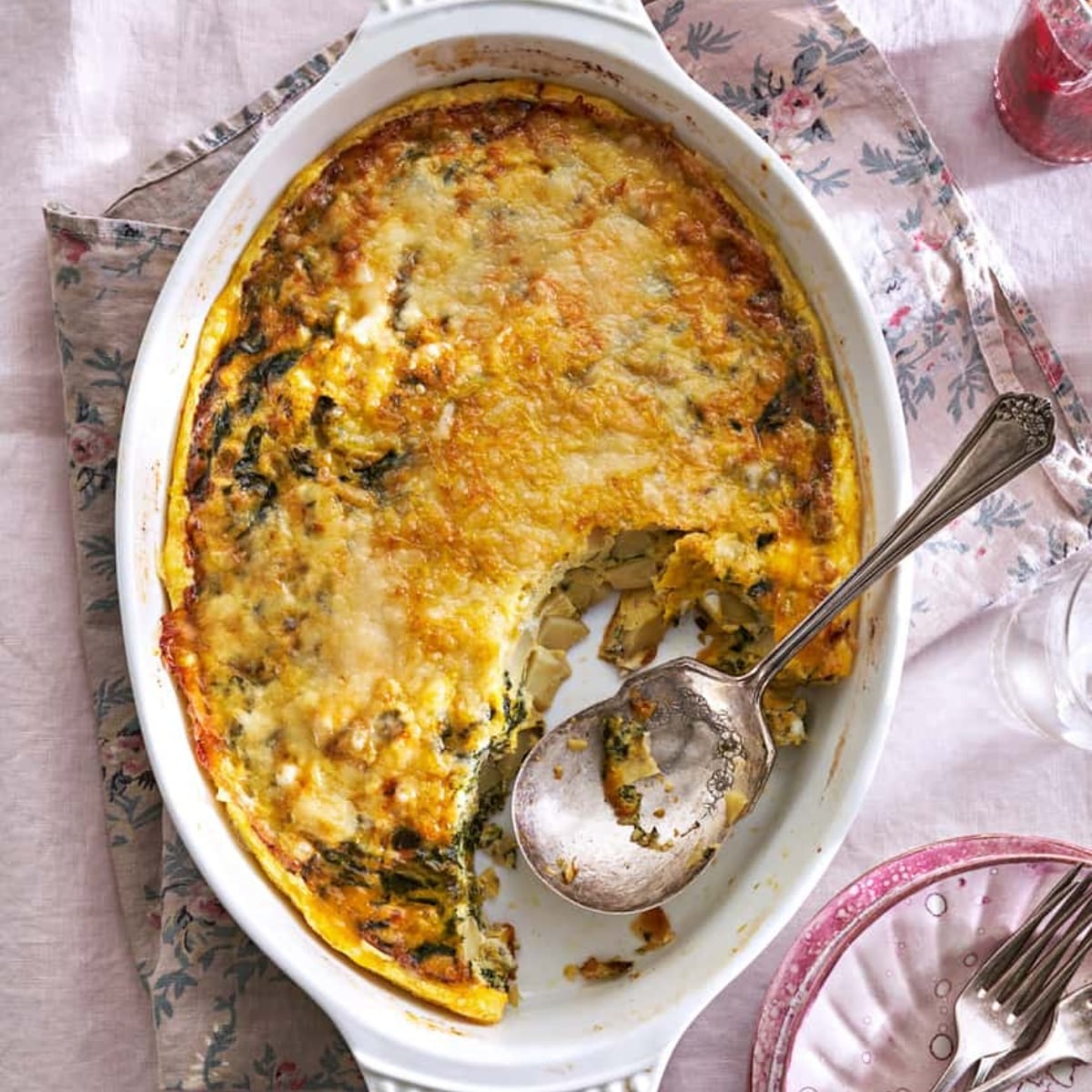 Oval baking dish with a partially served egg and vegetable casserole, perfect for Easter Breakfast recipes, a serving spoon inside, and remnants on a floral-patterned cloth and plate nearby.