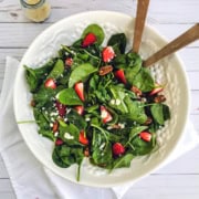 A white bowl of strawberry spinach salad with pecans and crumbled cheese, served with wooden tongs on a white napkin.
