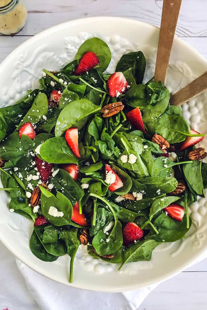A white bowl of strawberry spinach salad with pecans and crumbled cheese, served with wooden tongs on a white napkin.