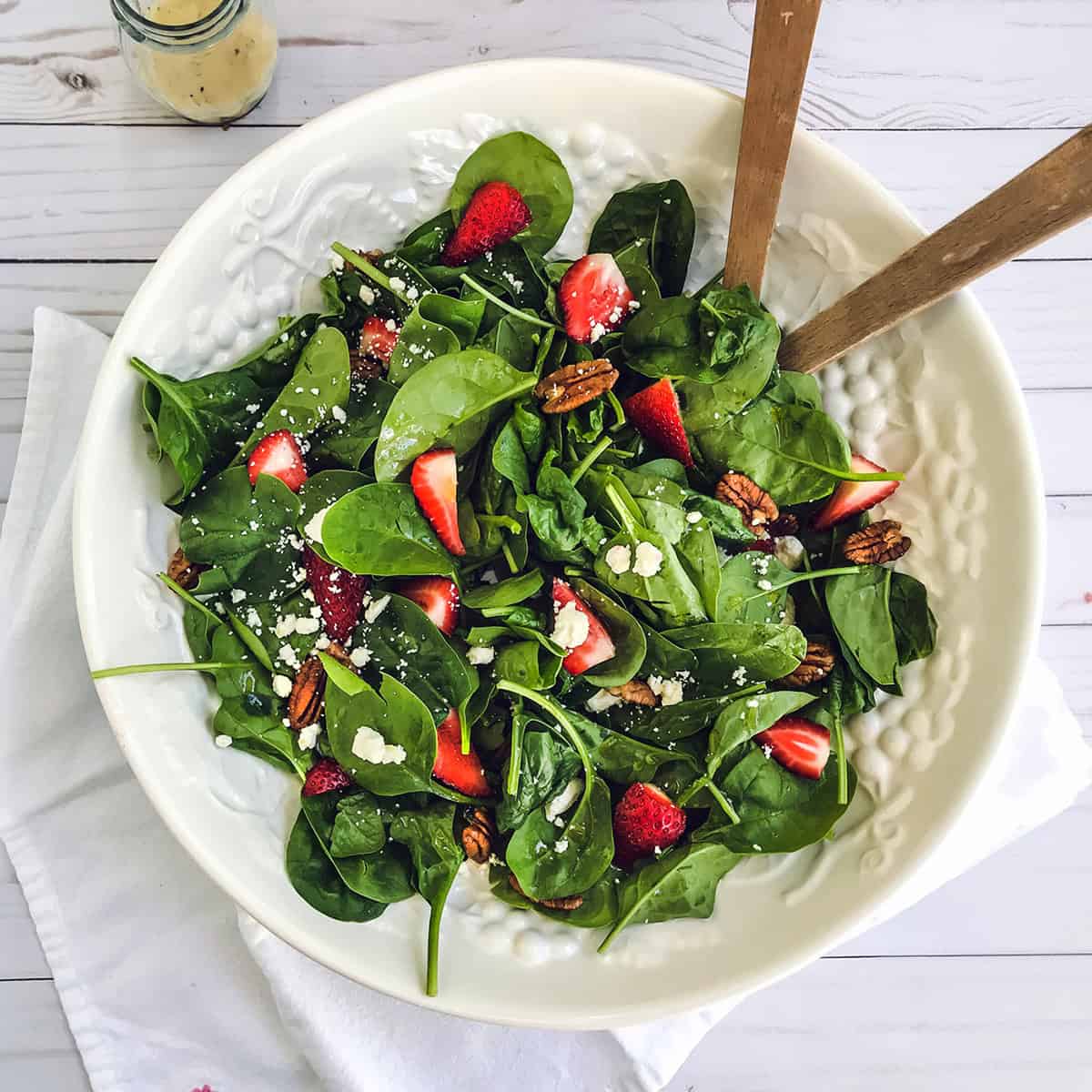 A white bowl of strawberry spinach salad with pecans and crumbled cheese, served with wooden tongs on a white napkin.