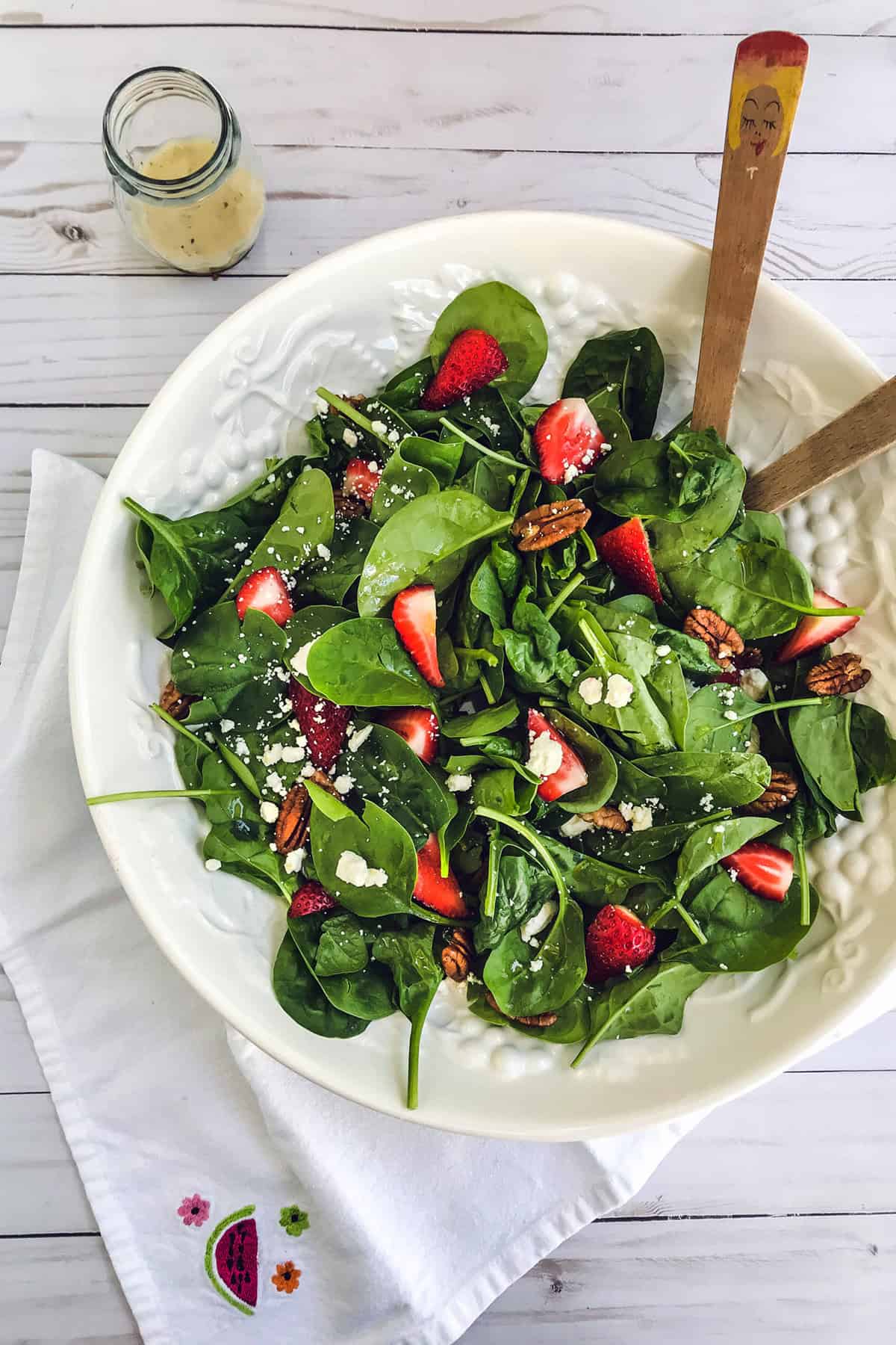 A white bowl filled with strawberry spinach salad-fresh spinach leaves, sliced strawberries, pecans, and crumbled feta cheese-with wooden salad servers, placed on a white napkin beside a small jar of dressing on a light wooden table.
