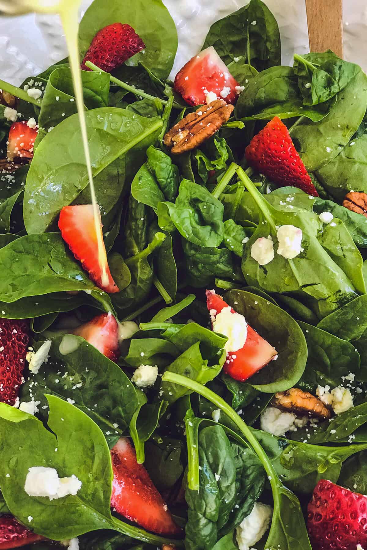 A close-up of a vibrant strawberry spinach salad with sliced strawberries, pecans, and crumbled feta cheese, as a stream of dressing is poured over the top.