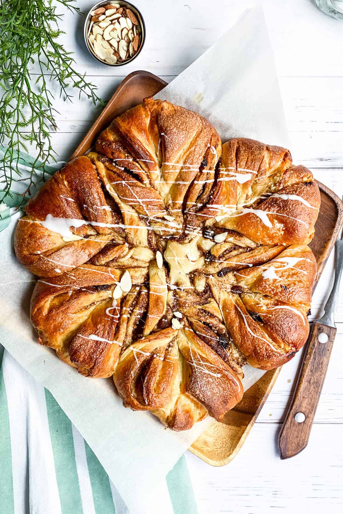 A star-shaped babka pastry with twisted segments, drizzled with white icing and sliced almonds, rests on parchment paper beside a wooden knife and a bowl of almonds.
