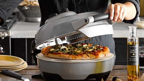 A person lifts the lid of a countertop pizza oven, revealing a cooked pizza—a perfect scene for father’s day gift ideas. Another pizza rests on a peel in the foreground with oil and utensils nearby.