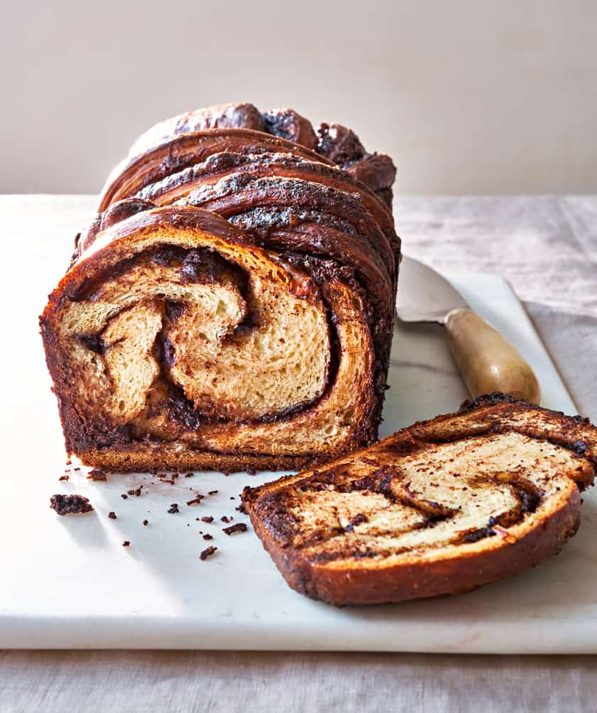 A loaf of chocolate babka with a swirl pattern, perfect for Easter Breakfast recipes, sits on a white cutting board with one slice cut and a knife beside it.