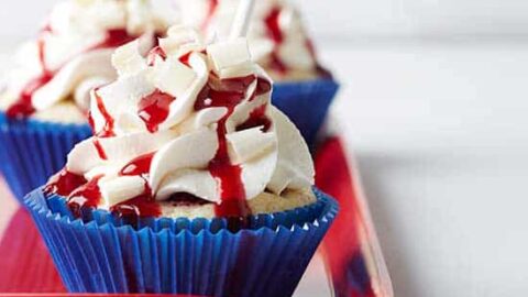 Cupcakes with white frosting, red syrup, and small blue star flags in blue wrappers are arranged on a red tray—perfect Memorial Day food for your celebration.