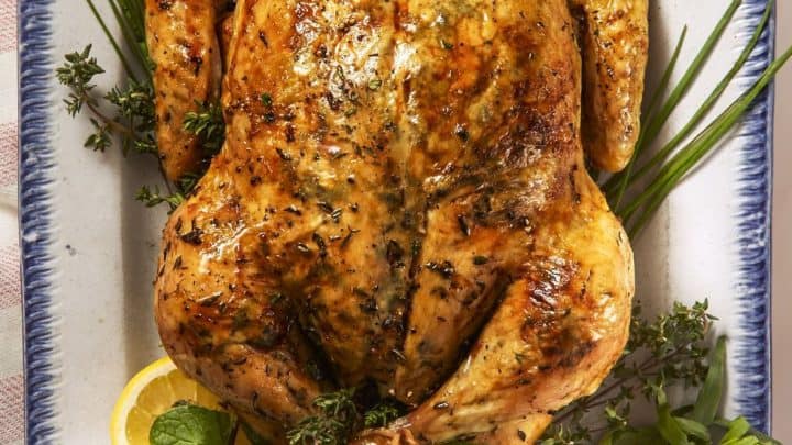 A whole roasted chicken garnished with fresh herbs and lemon halves, served on a white platter with a blue rim—a perfect centerpiece for Mother’s Day recipes.