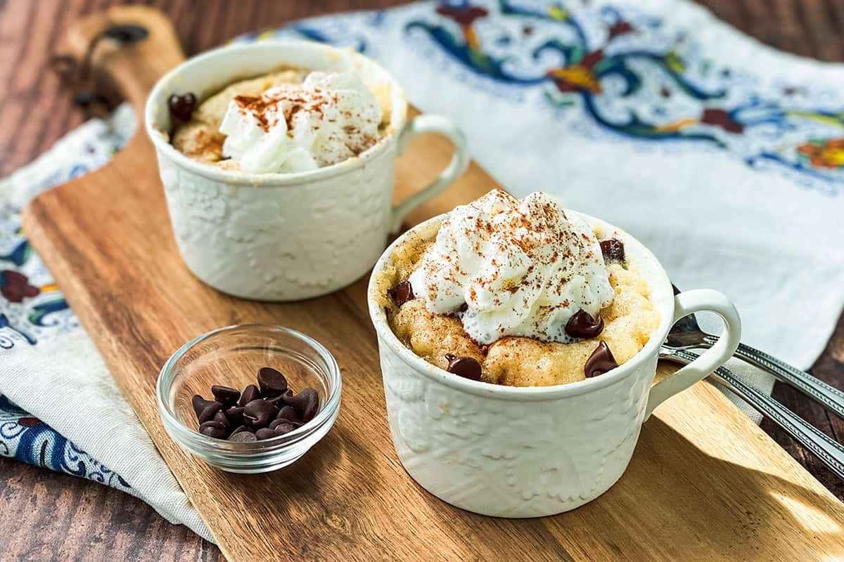 Two mugs with chocolate chip banana mug cakes topped with whipped cream and cinnamon sit on a wooden board, next to a small bowl of chocolate chips and a patterned cloth.