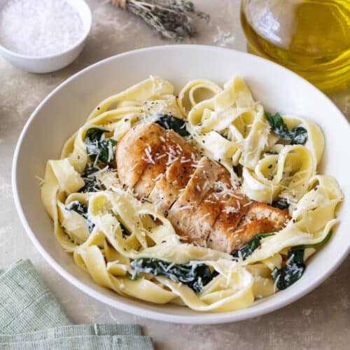 A white bowl filled with chicken fettuccine alfredo, spinach, and a grilled chicken breast topped with grated cheese. Surrounding the bowl are a fork, olive oil, salt, pepper, and a green napkin on a beige surface.