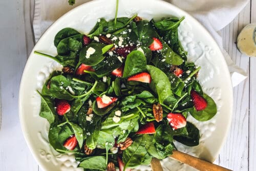 A white bowl filled with fresh strawberry spinach salad—sliced strawberries, spinach leaves, pecans, and crumbled cheese—with wooden salad utensils on the side. The bowl sits on a white table with a light napkin nearby.