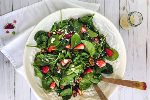 A bowl of strawberry spinach salad with sliced strawberries, pecans, and crumbled cheese sits on a white wooden table, surrounded by salad tongs, a watermelon napkin, and a small jar of dressing.