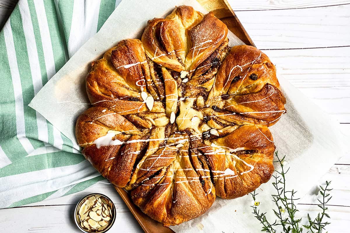 A round braided babka with a swirl pattern, drizzled with white icing, sits on parchment paper beside a bowl of sliced almonds and a striped green kitchen towel.