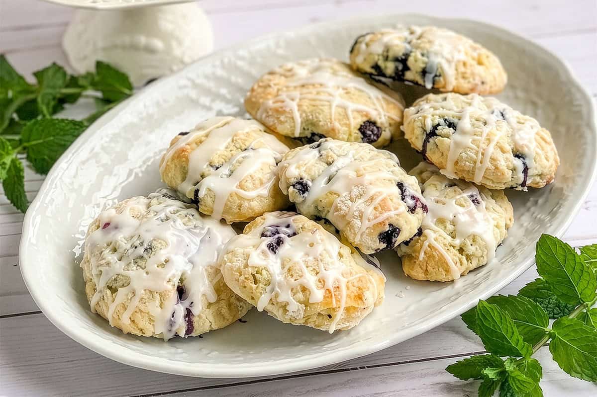A white plate with eight glazed blueberry scones, some with visible blueberries, is placed on a white wooden surface with green mint sprigs nearby.