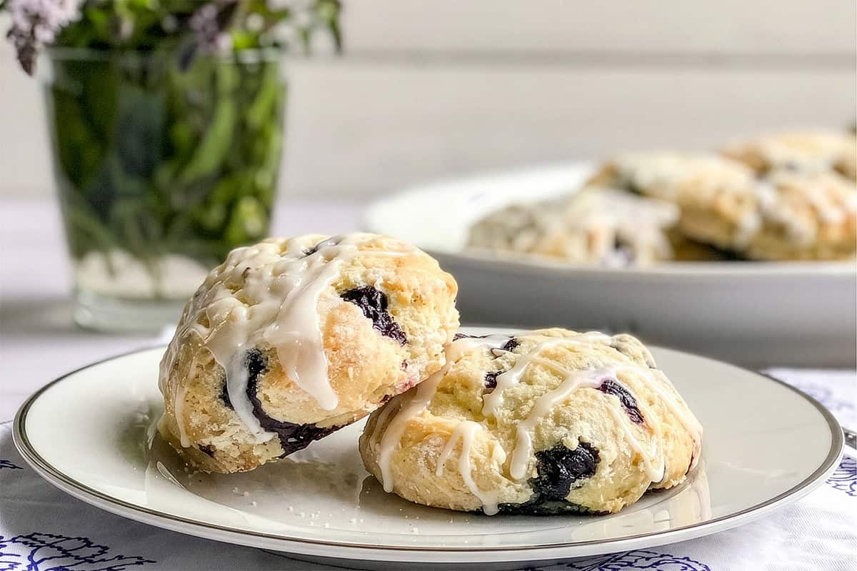 Two blueberry scones with sweet icing sit on a white plate, while more blueberry scones and a glass vase of green foliage create a soft, inviting backdrop.