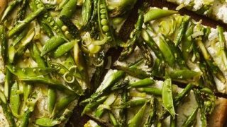 A sliced pizza topped with asparagus, snap peas, and cheese is displayed on a round wooden board—perfect for adding a fresh twist to your Mother’s Day recipes.