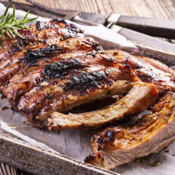 Cut grilled and seared back baby ribs on a wooden parchment lined serving tray with fresh rosemary on the side.