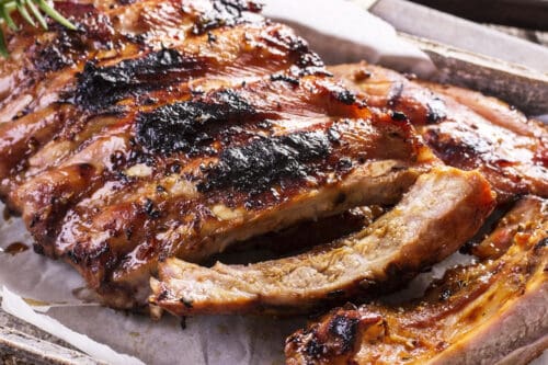Cut grilled and seared back baby ribs on a wooden parchment lined serving tray with fresh rosemary on the side.