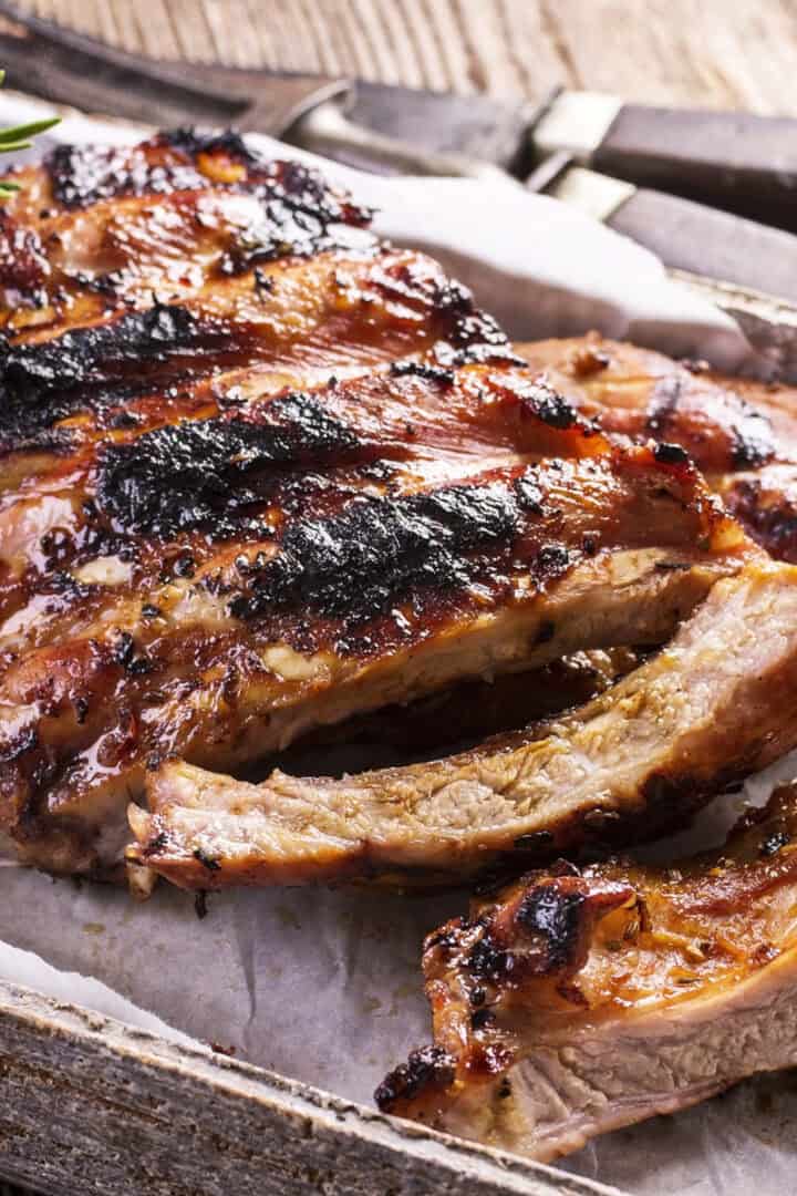 Cut grilled and seared back baby ribs on a wooden parchment lined serving tray with fresh rosemary on the side.