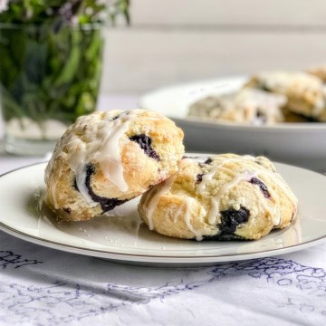 Two blueberry scones with white icing sit on a white plate, highlighting the delicious, homemade appeal of these blueberry scones. In the blurred background, a vase of greenery and more scones add to the inviting scene.