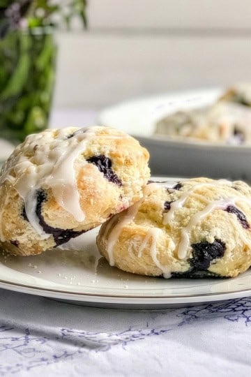 Two blueberry scones with white icing sit on a white plate, highlighting the delicious, homemade appeal of these blueberry scones. In the blurred background, a vase of greenery and more scones add to the inviting scene.