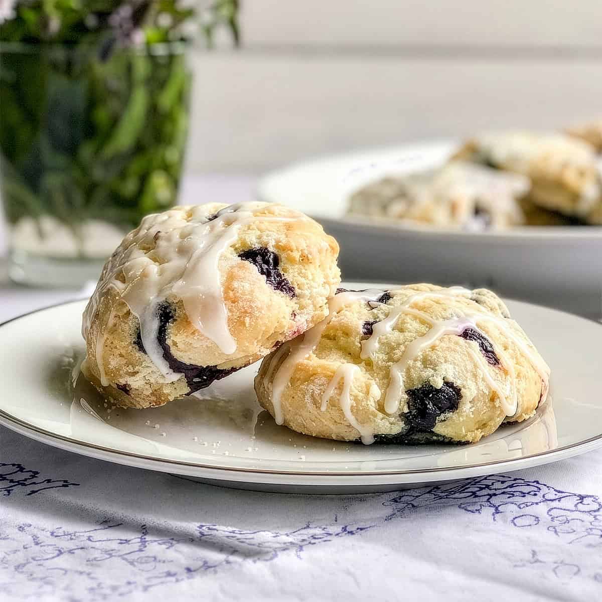 Two blueberry scones with white icing sit on a white plate, highlighting the delicious, homemade appeal of these blueberry scones. In the blurred background, a vase of greenery and more scones add to the inviting scene.