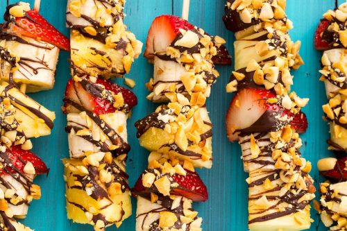 Four fruit kabobs with banana, strawberry, and pineapple pieces, drizzled with chocolate and sprinkled with chopped nuts, make colorful Memorial Day Desserts on wooden skewers over a bright blue wooden surface.