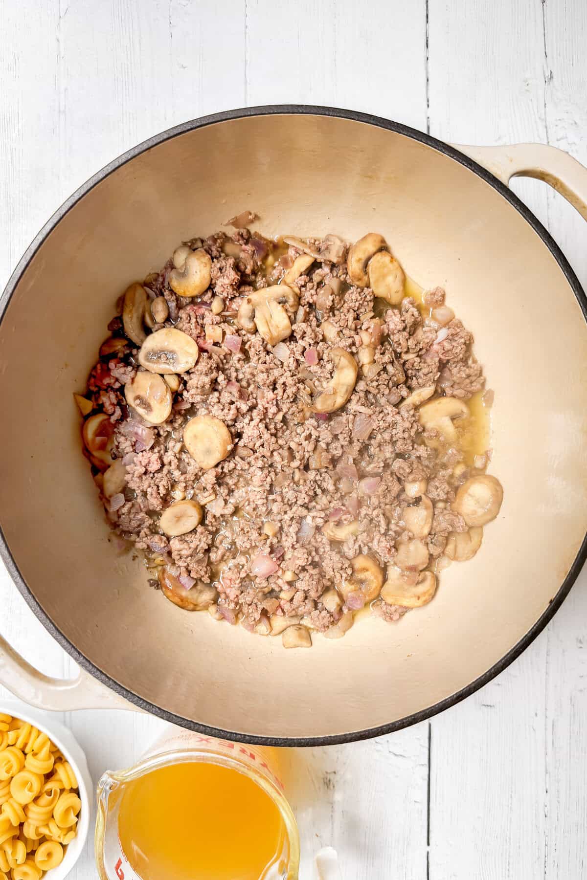 A large pot of cooked ground meat and sliced mushrooms sits on a white wooden surface, alongside uncooked pasta and a measuring cup of broth-perfect for making an easy One Pot Pasta meal.