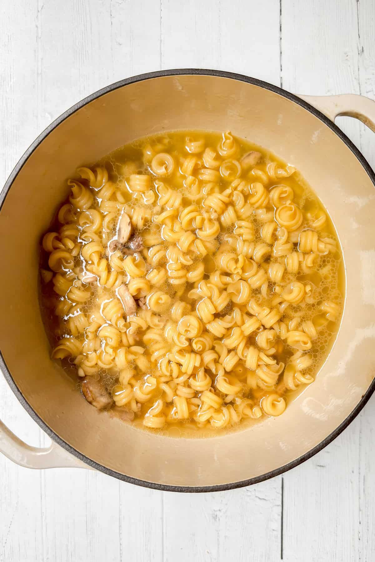 A large beige pot on a white wooden surface holds a comforting One Pot Pasta, featuring cooked noodles in a light, clear broth with tender pieces of chicken.