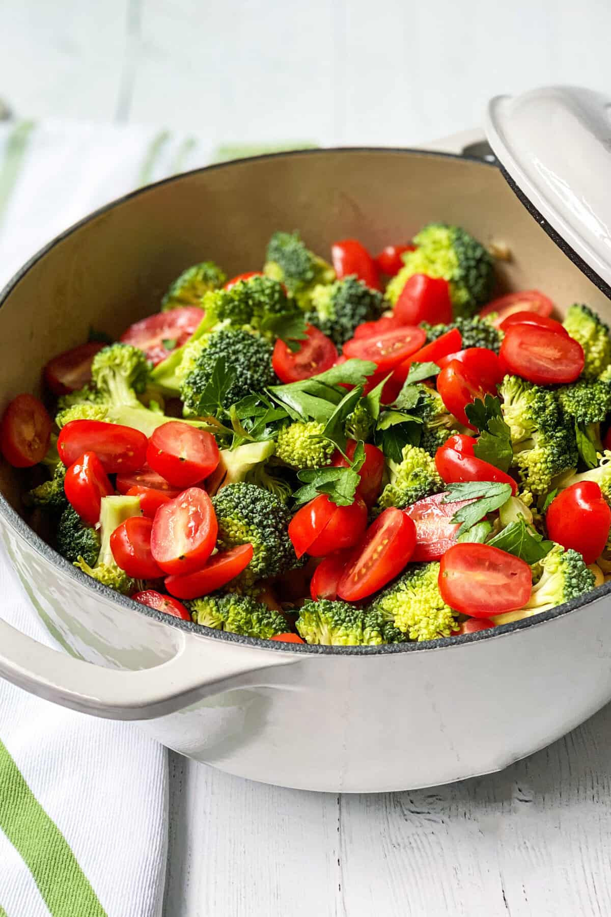 A white pot filled with fresh broccoli florets and halved cherry tomatoes, reminiscent of a delicious One Pot Pasta, is garnished with chopped parsley and sits on a white surface with a green-striped towel underneath.