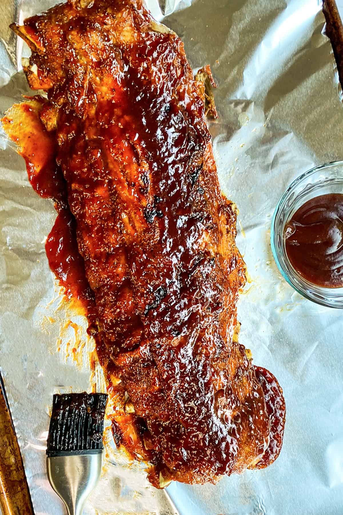 A rack of glazed barbecued ribs on foil with a basting brush and a small bowl of barbecue sauce beside it-these Easy Instant Pot Ribs are tender, juicy, and packed with flavor.
