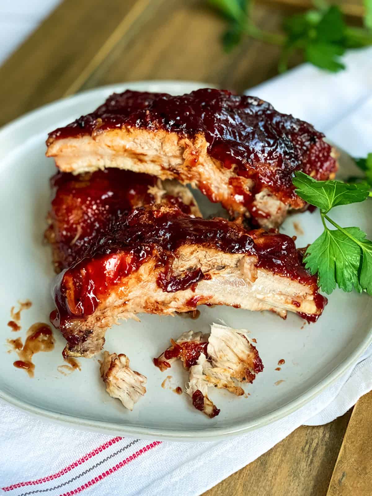 Three barbecue sauce-covered Easy Instant Pot Ribs on a white plate with a garnish of parsley, showing bite marks and pieces of meat on the plate.