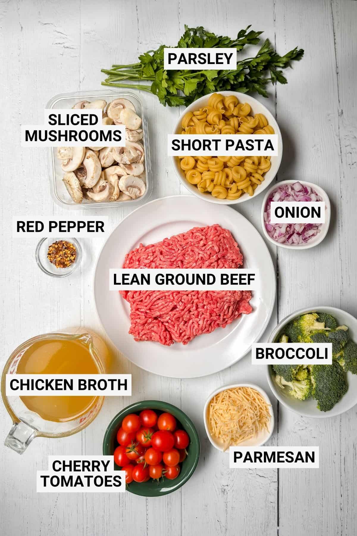 Top-down view of labeled One Pot Pasta ingredients on a white surface: parsley, sliced mushrooms, short pasta, onion, broccoli, lean ground beef, red pepper, chicken broth, cherry tomatoes, and parmesan.
