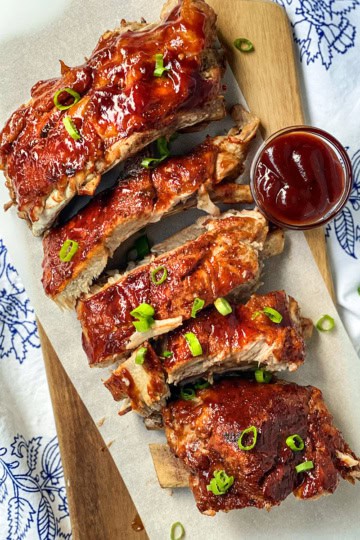 A stack of Easy Instant Pot Ribs glazed in barbecue sauce, garnished with chopped green onions, served on parchment paper with a small cup of barbecue sauce on the side.