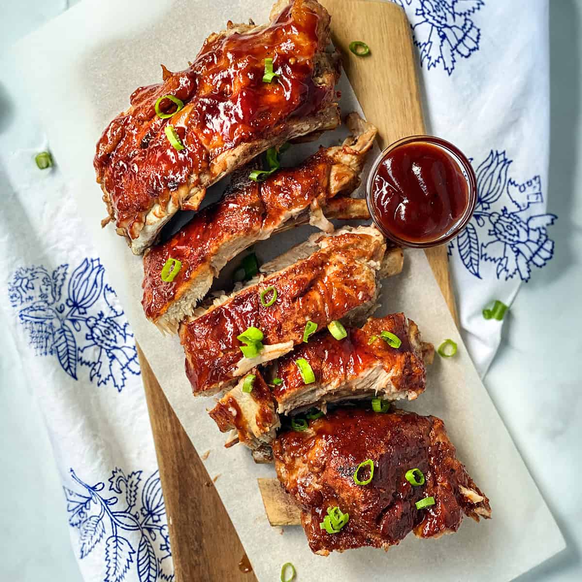 A stack of Easy Instant Pot Ribs glazed in barbecue sauce, garnished with chopped green onions, served on parchment paper with a small cup of barbecue sauce on the side.