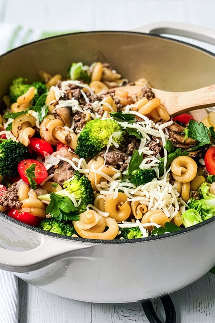 A pot of One Pot Pasta with broccoli, cherry tomatoes, ground meat, mushrooms, parsley, and shredded cheese, all mixed together with a wooden spoon inside.