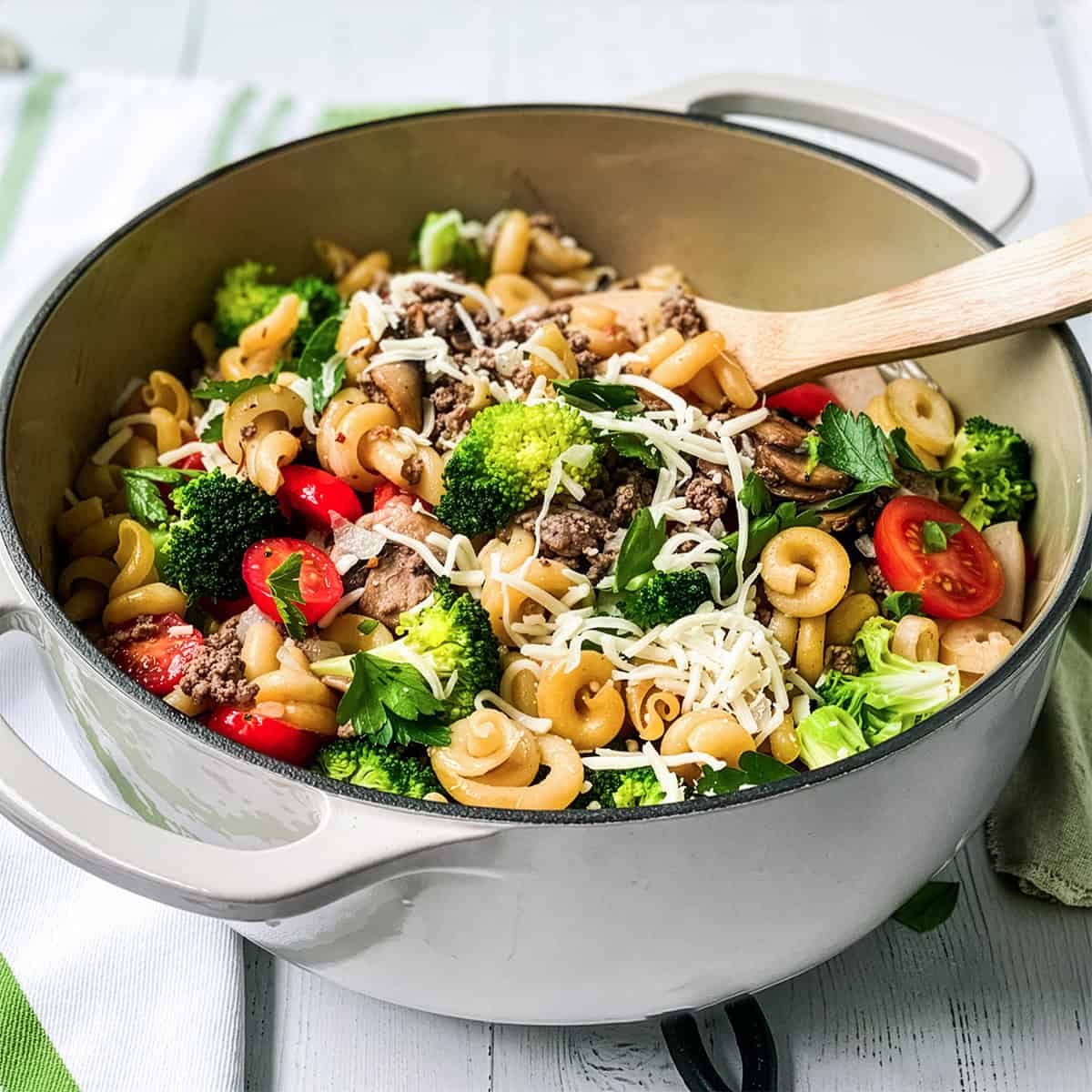 A pot of One Pot Pasta with broccoli, cherry tomatoes, ground meat, mushrooms, parsley, and shredded cheese, all mixed together with a wooden spoon inside.