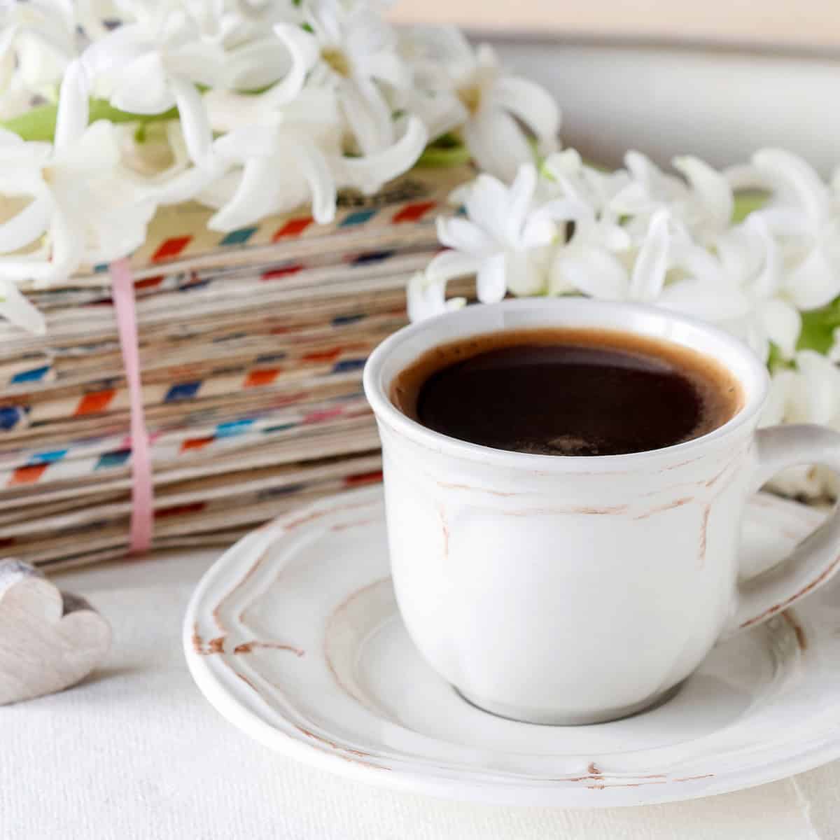 A white cup of black coffee sits on a saucer beside a stack of old letters tied with string, with white flowers in the background—perfect for reflecting on inspiring Mother's Day messages.
