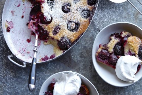 An oval baking dish of cherry clafoutis with a serving spoon, two bowls with clafoutis and whipped cream, and a sifter with powdered sugar on a gray surface-perfect for Memorial Day Desserts.