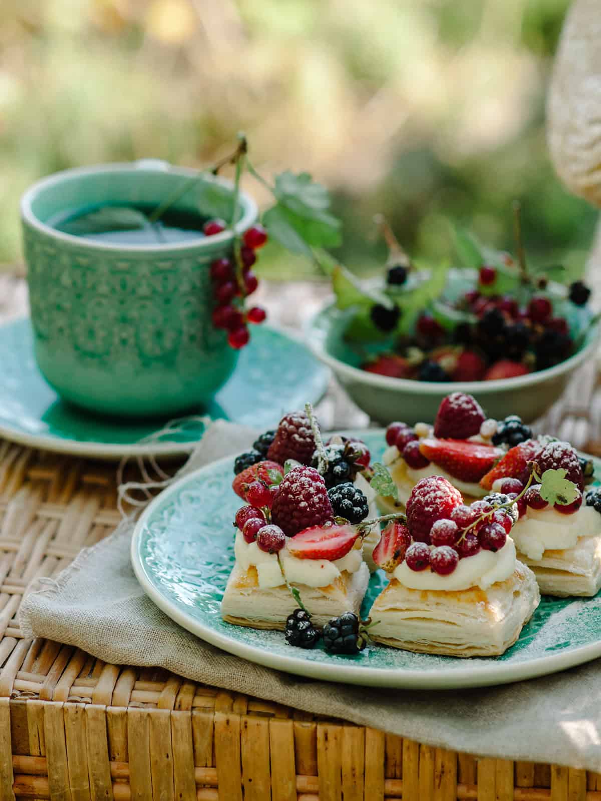 A turquoise cup of tea and a plate with puff pastry tarts—delicate squares topped with cream and assorted berries—sit on a wicker tray outdoors.