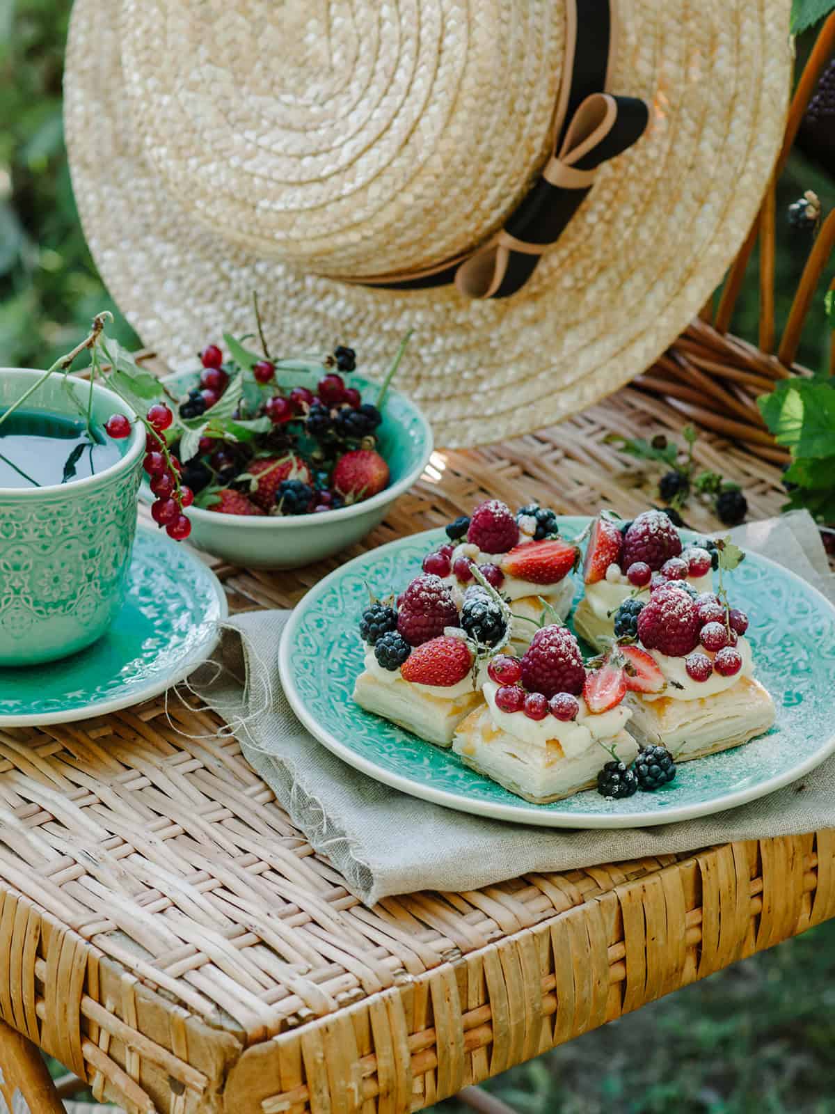 A wicker chair holds a plate of puff pastry tarts topped with berries, a bowl of mixed berries, a patterned cup and saucer, and a straw hat with a black ribbon.