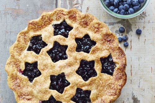 A lattice-topped blueberry pie sits on a rustic wooden surface next to a small glass bowl filled with fresh blueberries, making it a perfect centerpiece for Memorial Day Desserts.