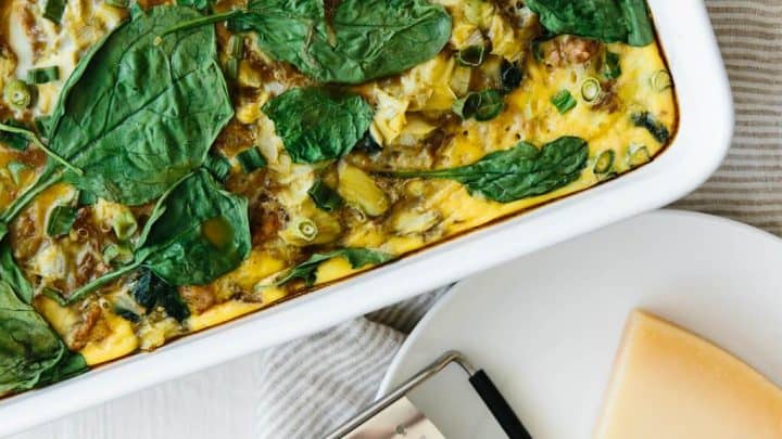 A baked egg casserole with spinach in a white dish sits next to a plate holding a wedge of cheese and a cheese slicer—perfect inspiration for breakfast casserole recipes.