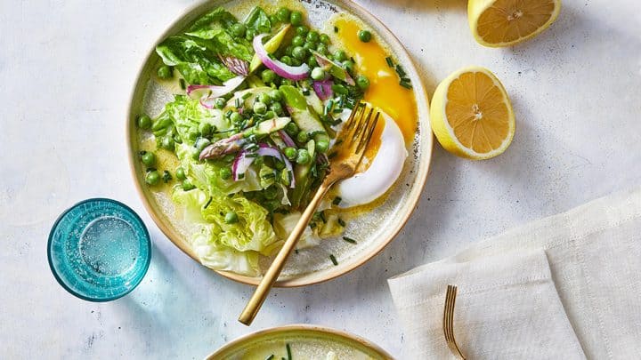 Three plates of salad with lettuce, peas, asparagus, poached eggs, and red onion slices, served with lemon halves and two blue glasses of water on a white surface—perfect for light Mother’s Day recipes.