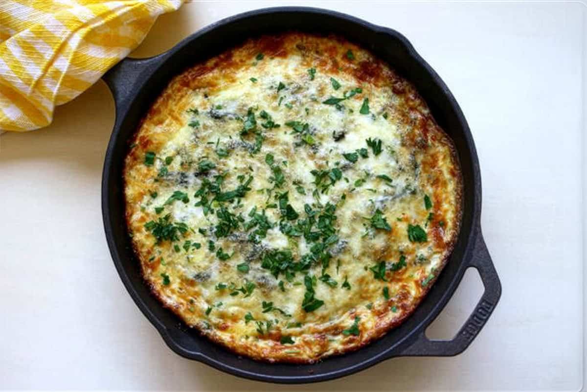 A ham and roasted asparagus frittata with herbs on top sits in a black cast iron skillet on a light surface, next to a yellow and white checkered cloth.