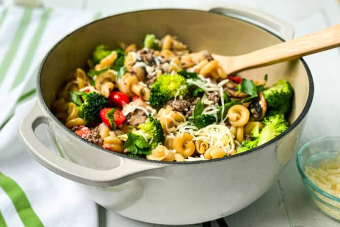 A large pot filled with One Pot Pasta, ground meat, broccoli, cherry tomatoes, mushrooms, and melted cheese, with a wooden spoon resting inside. A small bowl of grated cheese sits nearby on a white surface.
