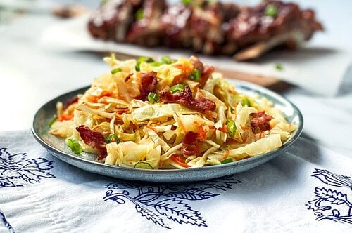 A plate of hot slaw featuring cabbage salad with carrots and bacon bits, garnished with green onions. The dish rests on a patterned cloth, with a blurred view of ribs in the background on a wooden board.