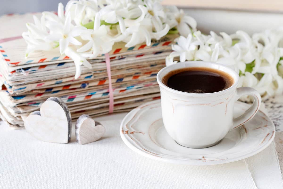 A white cup of black coffee on a saucer sits next to stacked letters, wooden heart shapes, and white flowers-perfect inspiration for crafting inspiring Mother's Day messages.